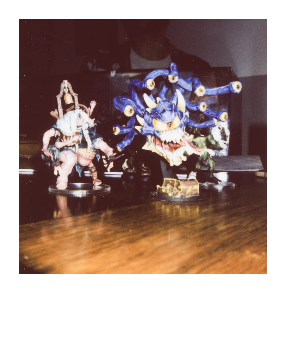 Painted fantasy RPG miniatures, including a large eye-covered monster, displayed on a wooden table.